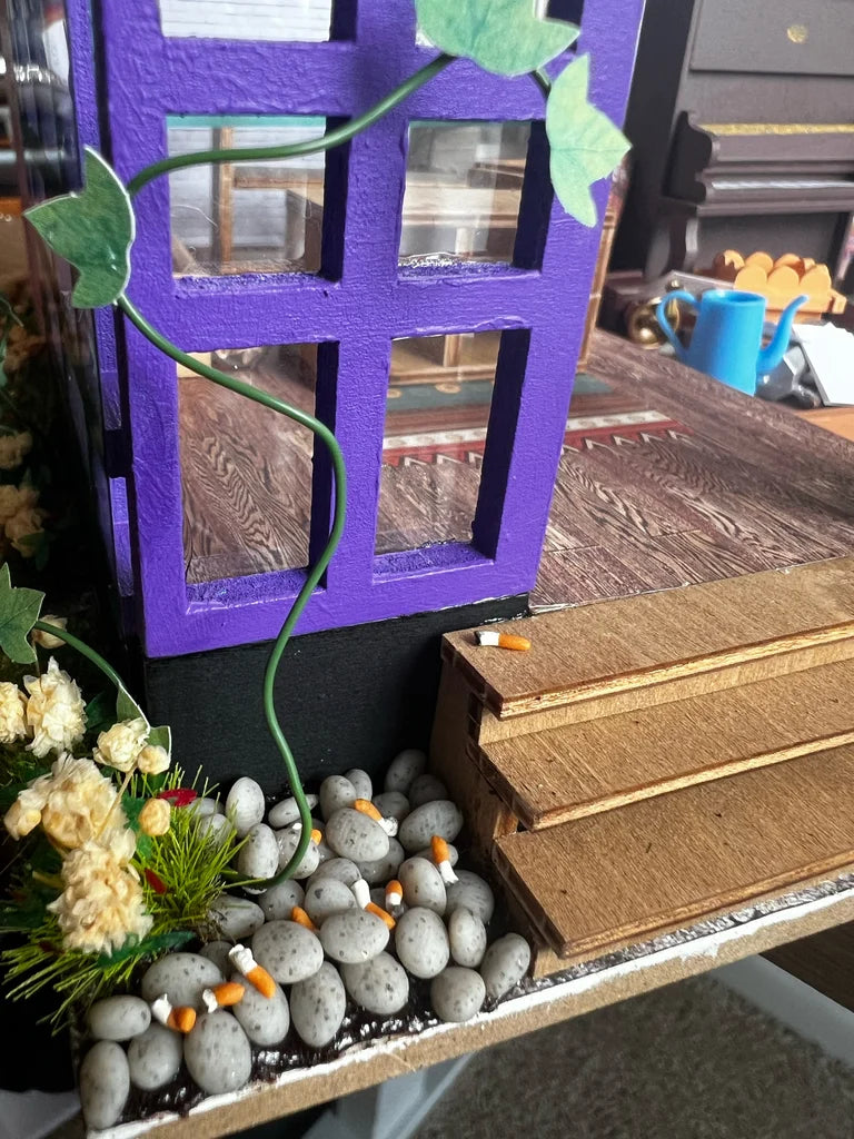 Miniature garden with purple window, stones, and wooden steps on a table.