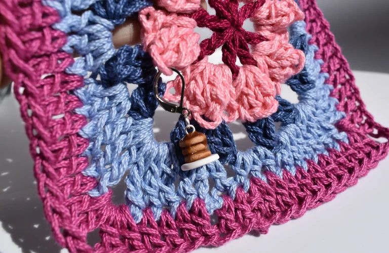 Crocheted square with pink and blue patterns and a pancake charm on a white background