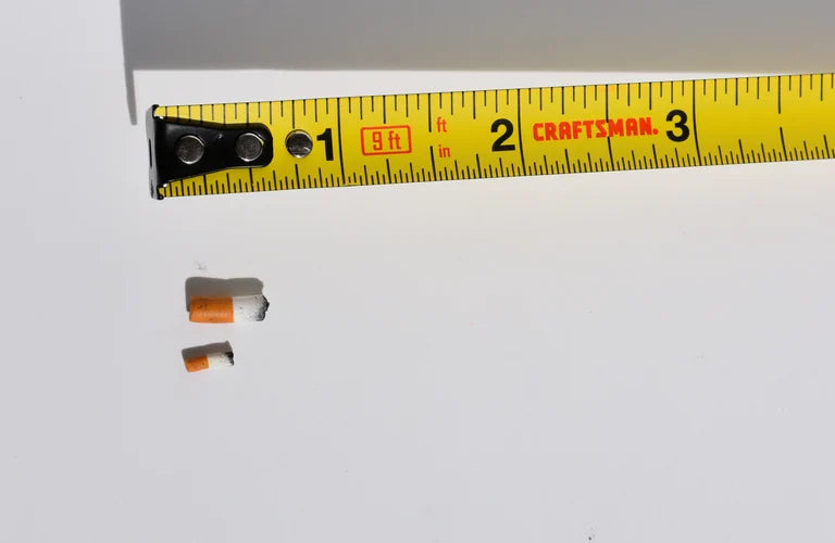 Two small miniature handmade cigarettes next to a yellow Craftsman tape measure on a white background one is 1:12 scale and the other is 1:6 scale.