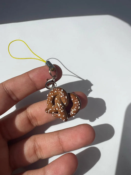 Hand holding a small gold pretzel-shaped keychain with a yellow string on a white surface.