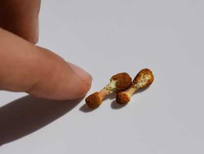 Two miniature fried brown chicken wings, both drumsticks. They are positioned next to a human figure for scale, on a white background.