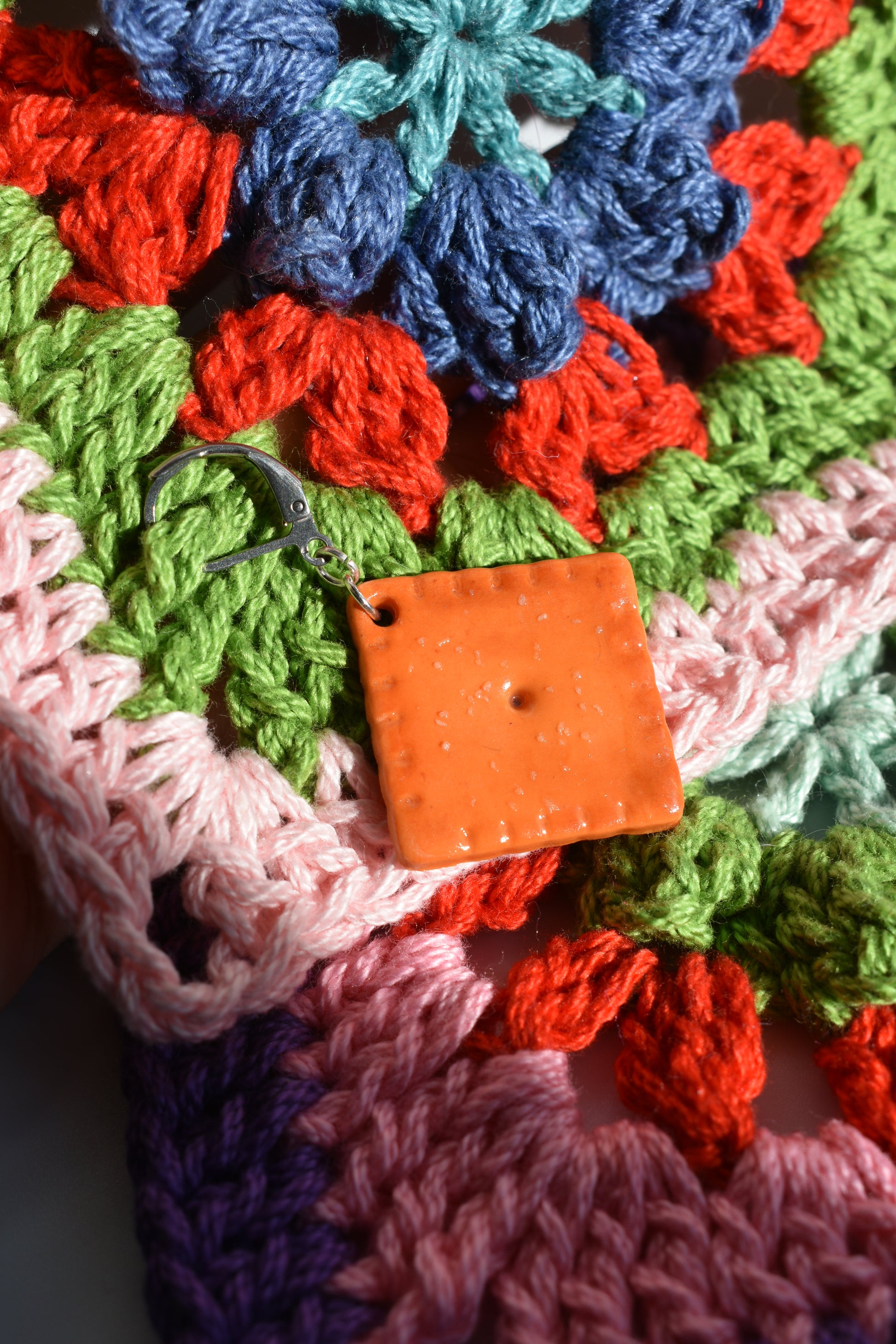 Colorful crocheted fabric with a crochet marker holding stitches together. The square is orange and made to look like a cheddar cheese cracker in the center of the image. 