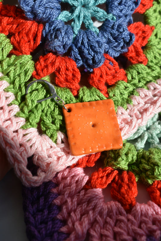 Colorful crocheted fabric with a crochet marker holding stitches together. The square is orange and made to look like a cheddar cheese cracker in the center of the image. 