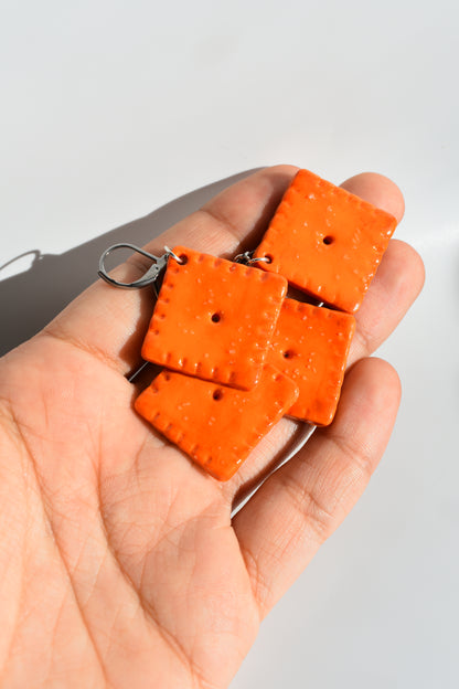 Hand holding four orange square cheddar cracker looking earrings with a plain white background