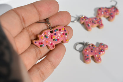 Hand holding a pink animal cookie-shaped keychain with sprinkles, with more keychains in the background.