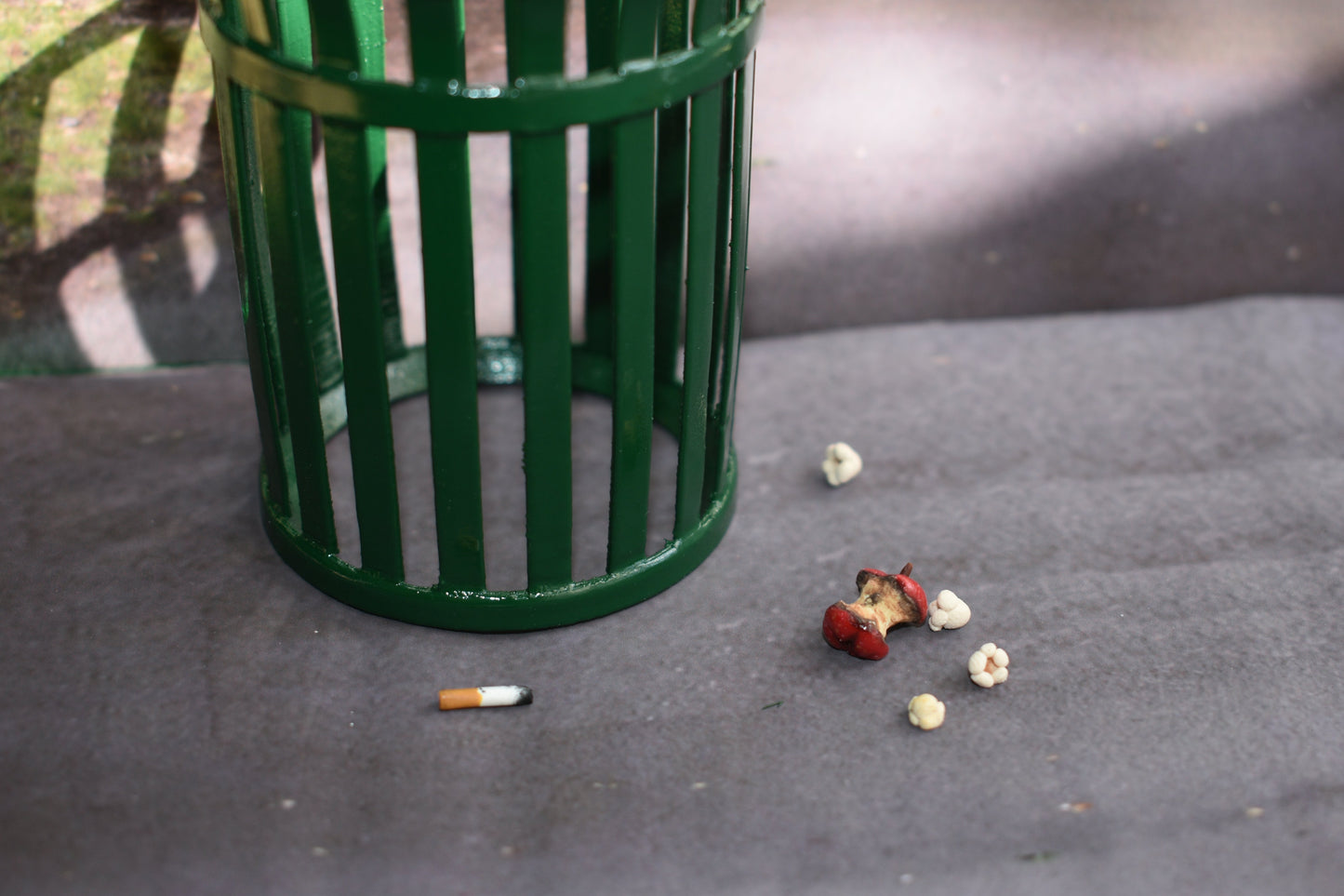 Miniature green trash can on a concrete surface with scattered trash items nearby. Items are 4 pieces of miniature popcorn, a red apple core that is rotting, and a singular amber and white cigarette butt with burnt tip.