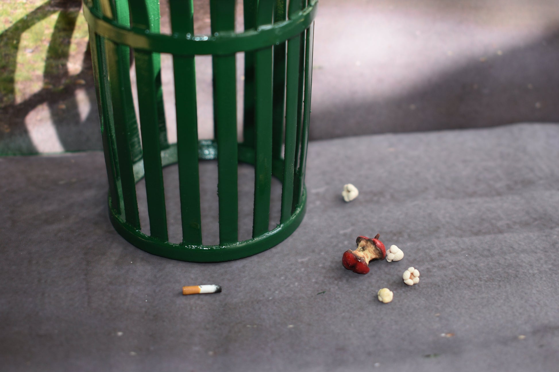 Miniature green trash can on a concrete surface with scattered trash items nearby. Items are 4 pieces of miniature popcorn, a red apple core that is rotting, and a singular amber and white cigarette butt with burnt tip.