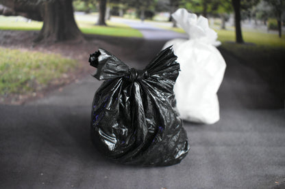 One black trash bag and one white trash bag on a sidewalk with a blurred park background