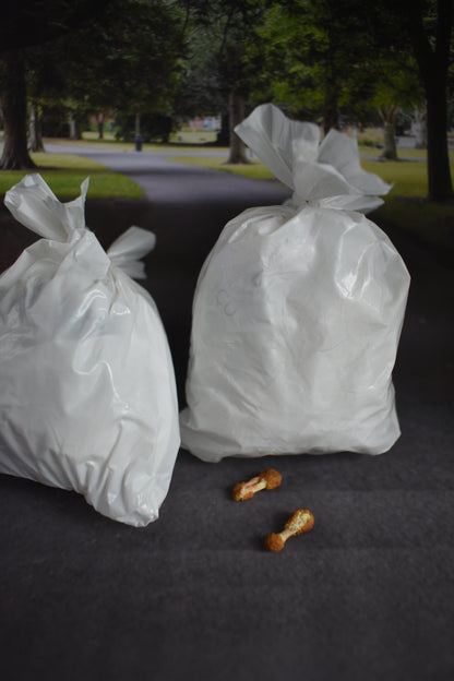 Two white plastic trash bags on a dark asphalt-like surface, blurred park background and two discarded chicken wings.