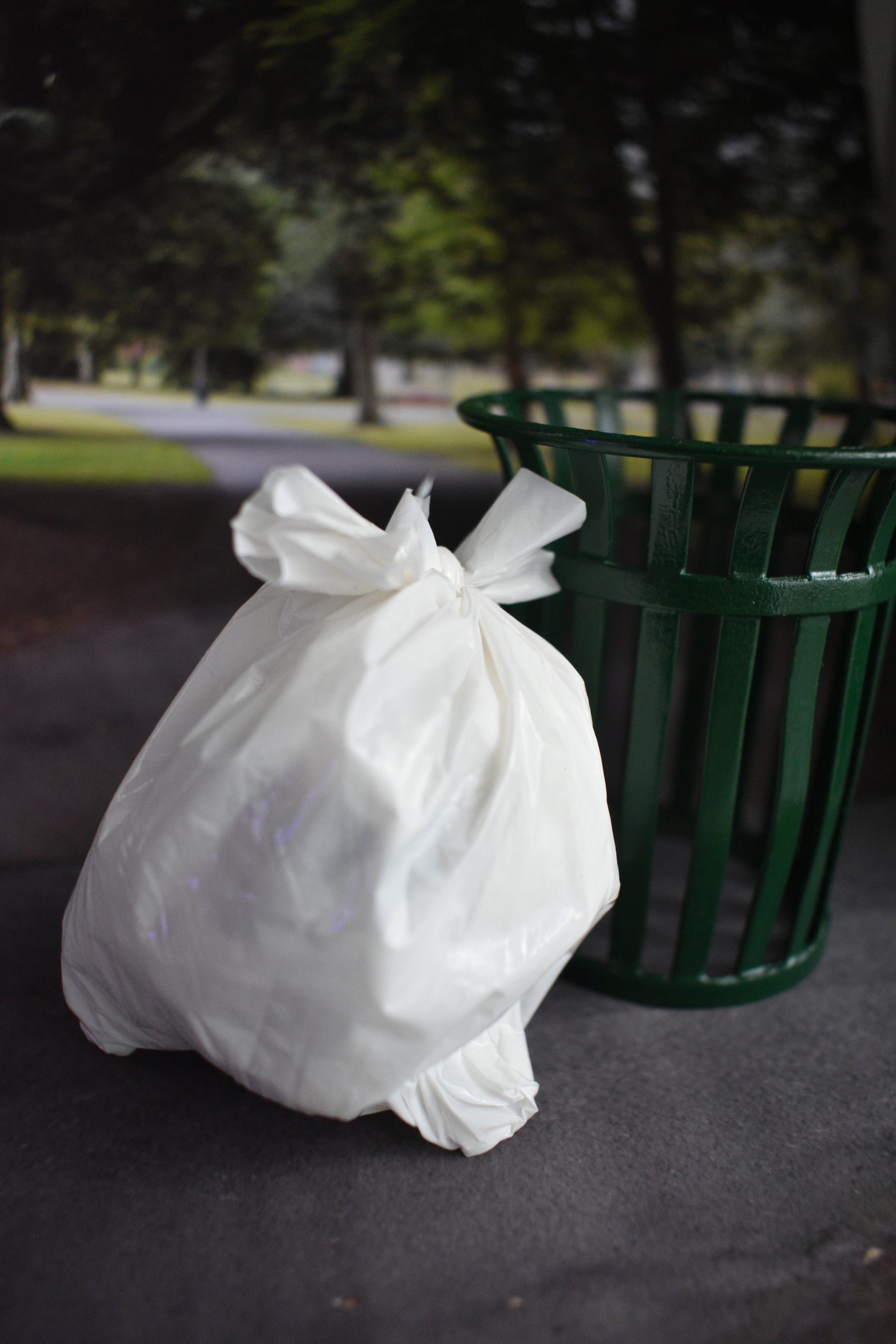 Miniature white plastic trash bag on a dark, asphalt-like surface with a green trash can in the background