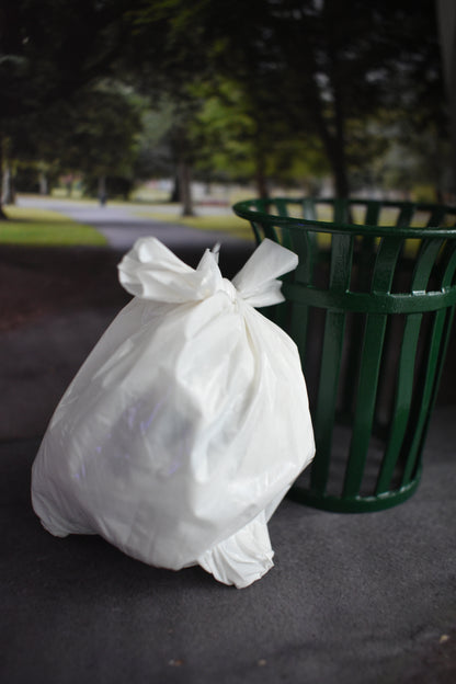 Miniature white plastic trash bag on a dark, asphalt-like surface with a green trash can in the background