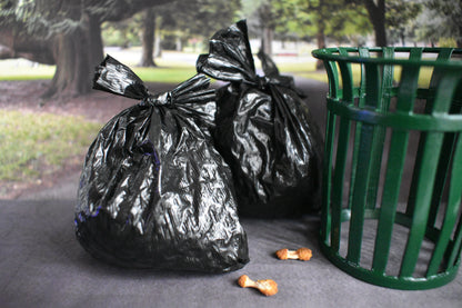 Two miniature black trash bags next to a green trash can in a park setting. At the base of the trash bags there are two discarded realistic chicken wings. One is a drum stick and one is a flat.