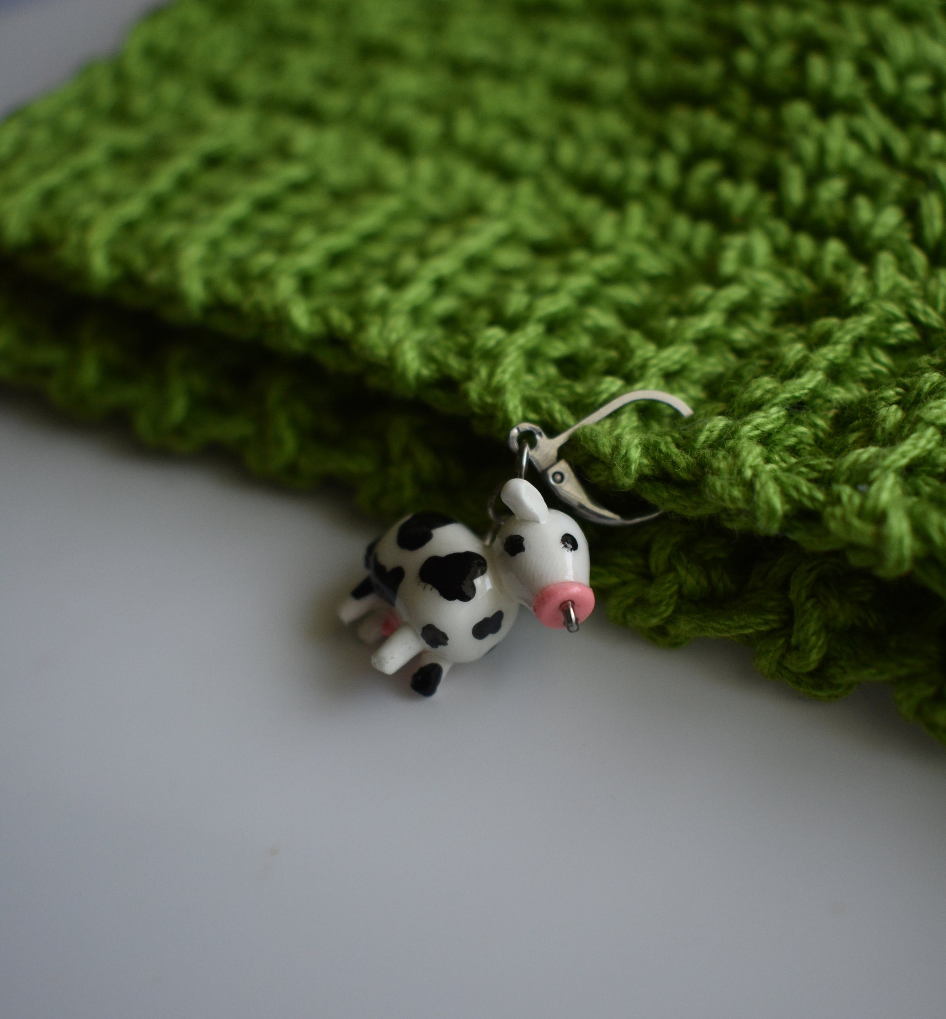 A small cow-shaped charm with black and white spots and a pink nose hangs from a silver wire loop, resting on a folded bright green knitted fabric against a light gray background.