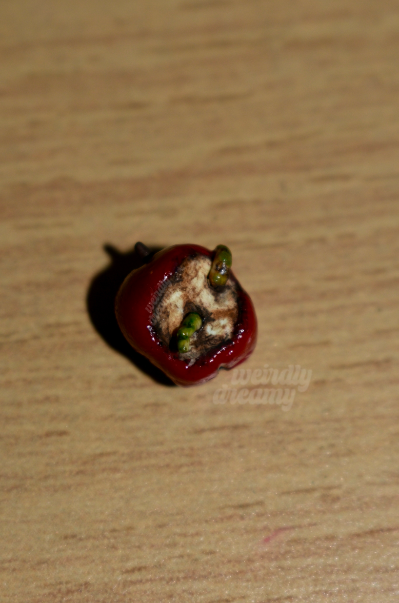 Miniature red apple with two green slimy worms working their way out of the center of the oxidized apple core on a wooden surface
