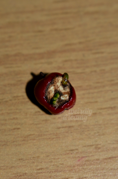 Miniature red apple with two green slimy worms working their way out of the center of the oxidized apple core on a wooden surface