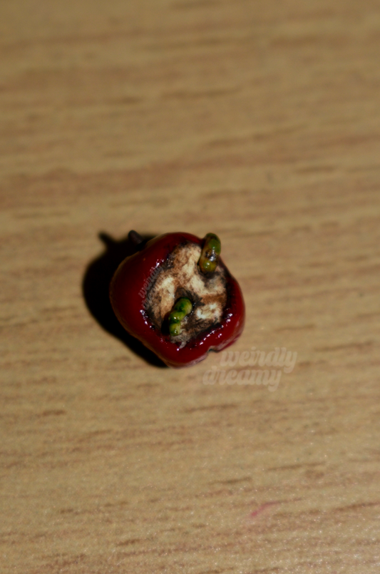 Miniature red apple with two green slimy worms working their way out of the center of the oxidized apple core on a wooden surface