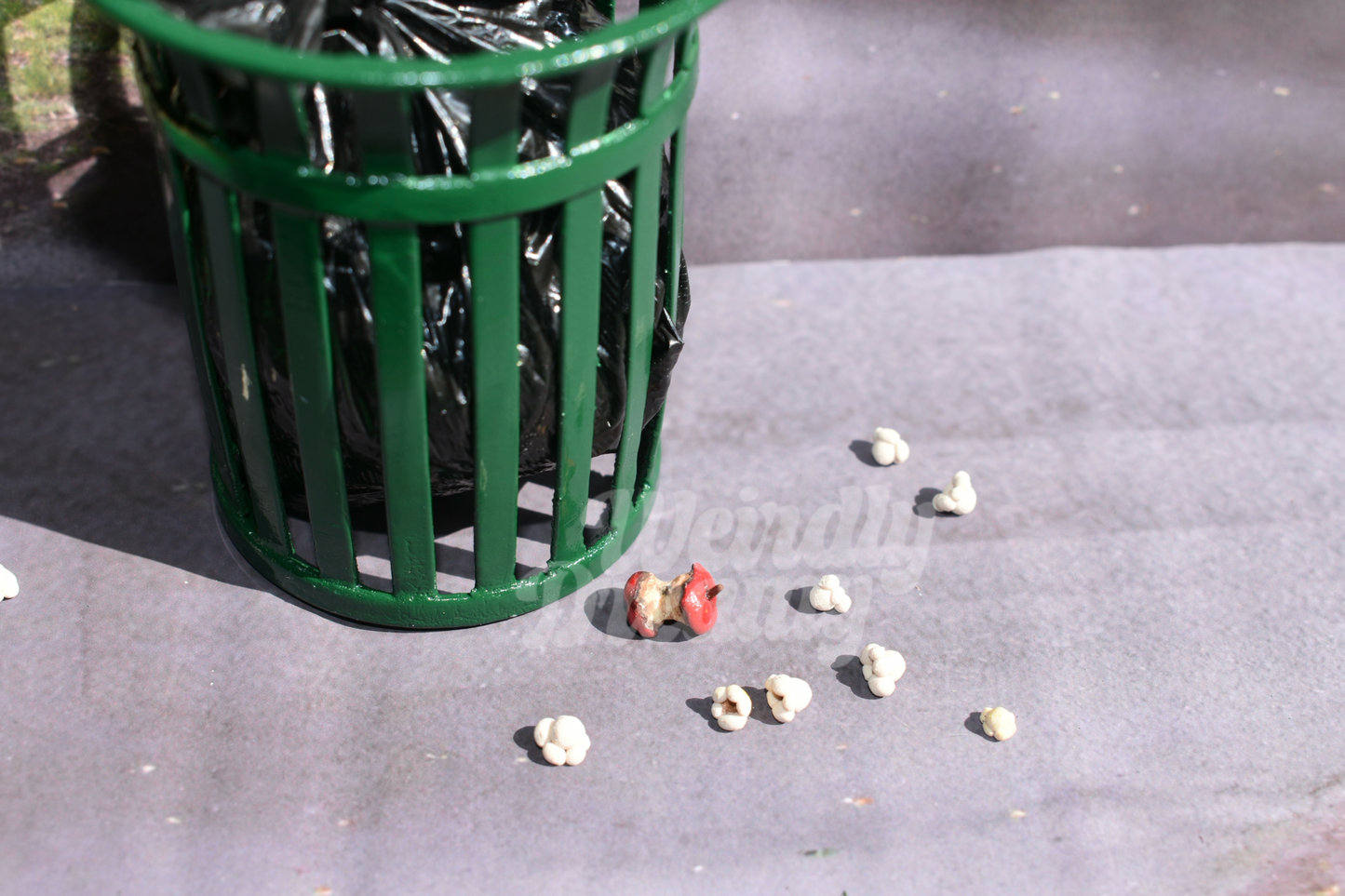 Green trash can with black trash bag liner. Scattered at the base of trash can on the asphalt are 8 pieces of miniature realistic popcorn and a singular miniature red apple core. The apple is oxidized and rotting. 