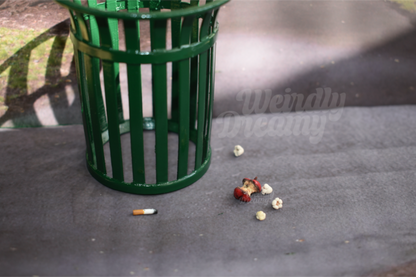 Miniature green trash can on a concrete surface with scattered trash items nearby. Items are 4 pieces of miniature popcorn, a red apple core that is rotting, and a singular amber and white cigarette butt with burnt tip.