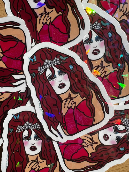 Sticker of a stylized woman with red hair and a tiara on a wooden surface.