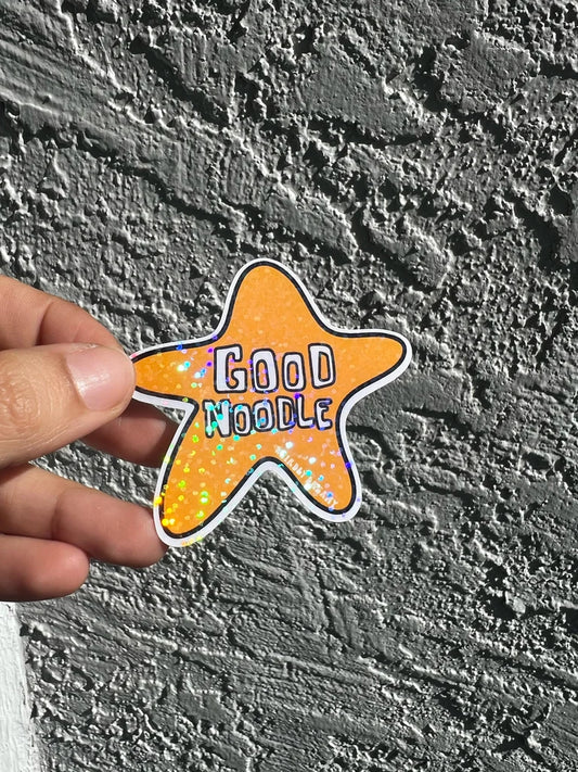 Star-shaped 'Good Noodle' sticker held against a textured gray wall.