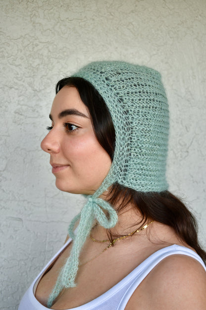 Woman wearing a light green knitted bonnet against a textured white background