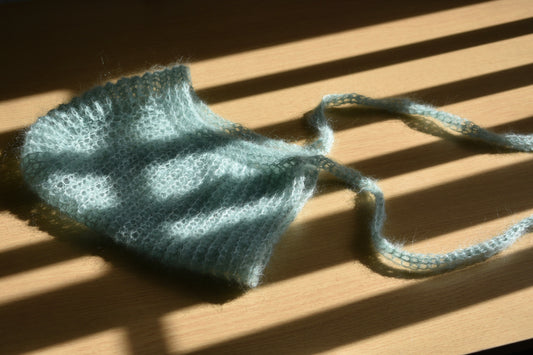 Knitted sage mohair silk crochet bonnet on a wooden surface with shadows