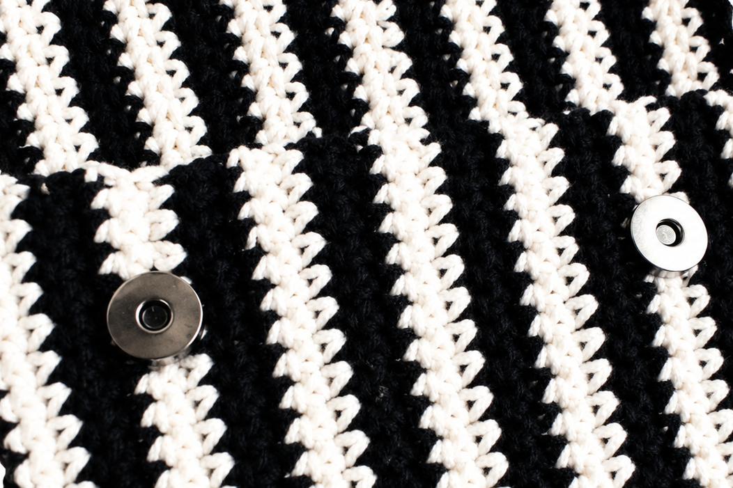 Close-up of a black and white striped crocheted pouch with metal snap buttons on a white background.